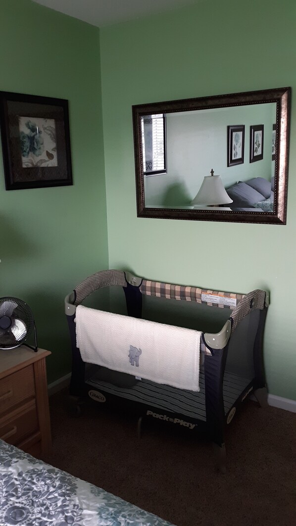 Une pièce avec un mur vert, un grand miroir, un berceau recouvert d’une couverture à carreaux, une table de chevet avec un ventilateur et une couvre-lit à motifs floraux.