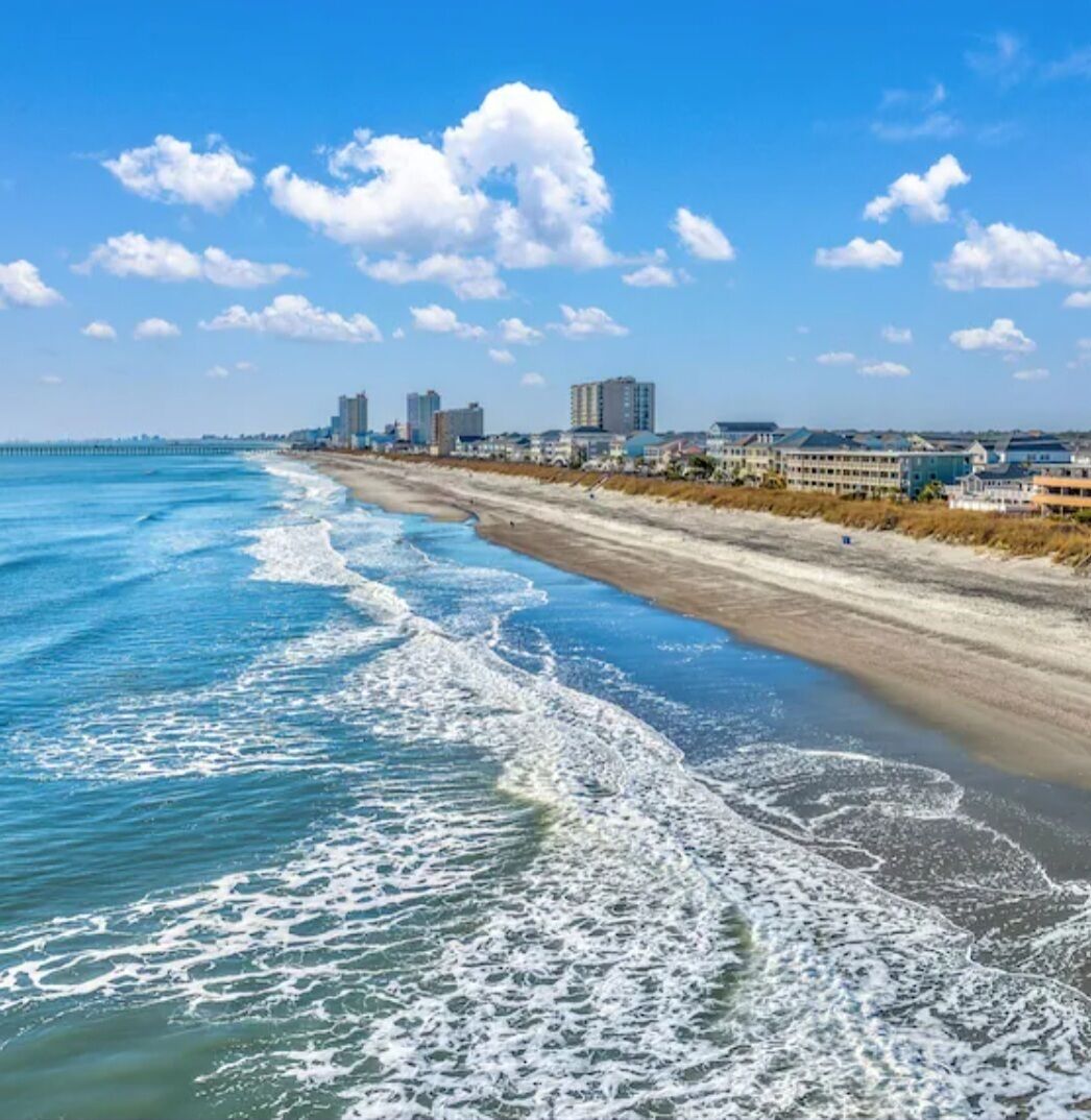 A beach with waves, a pier, and buildings in the background.