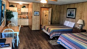 A compact room with two beds, a small kitchenette, a dining table, and a ceiling fan.