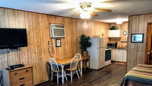 A compact room with a wooden wall, a ceiling fan, a TV, a refrigerator, a microwave, a dining table with chairs, and a bed with a striped blanket.
