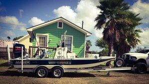 A boat trailer with a boat named 'MAGNUM' and 'CRAB CLAWSHER II' on it, parked in front of a green house with a white truck nearby.
