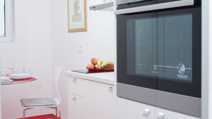 Une cuisine équipée d’un four moderne, un coin repas avec une chaise, et une table dressée avec des assiettes et un saladier rempli de fruits.