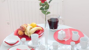Une table dressée pour deux personnes, avec une rose rouge dans un vase transparent, un panier de fruits, une carafe de boisson rouge et de la vaisselle blanche.