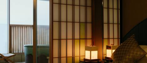 A hotel room with a bed, a nightstand with a lamp, a sliding shoji screen, a window with blinds, and a view of the sea.