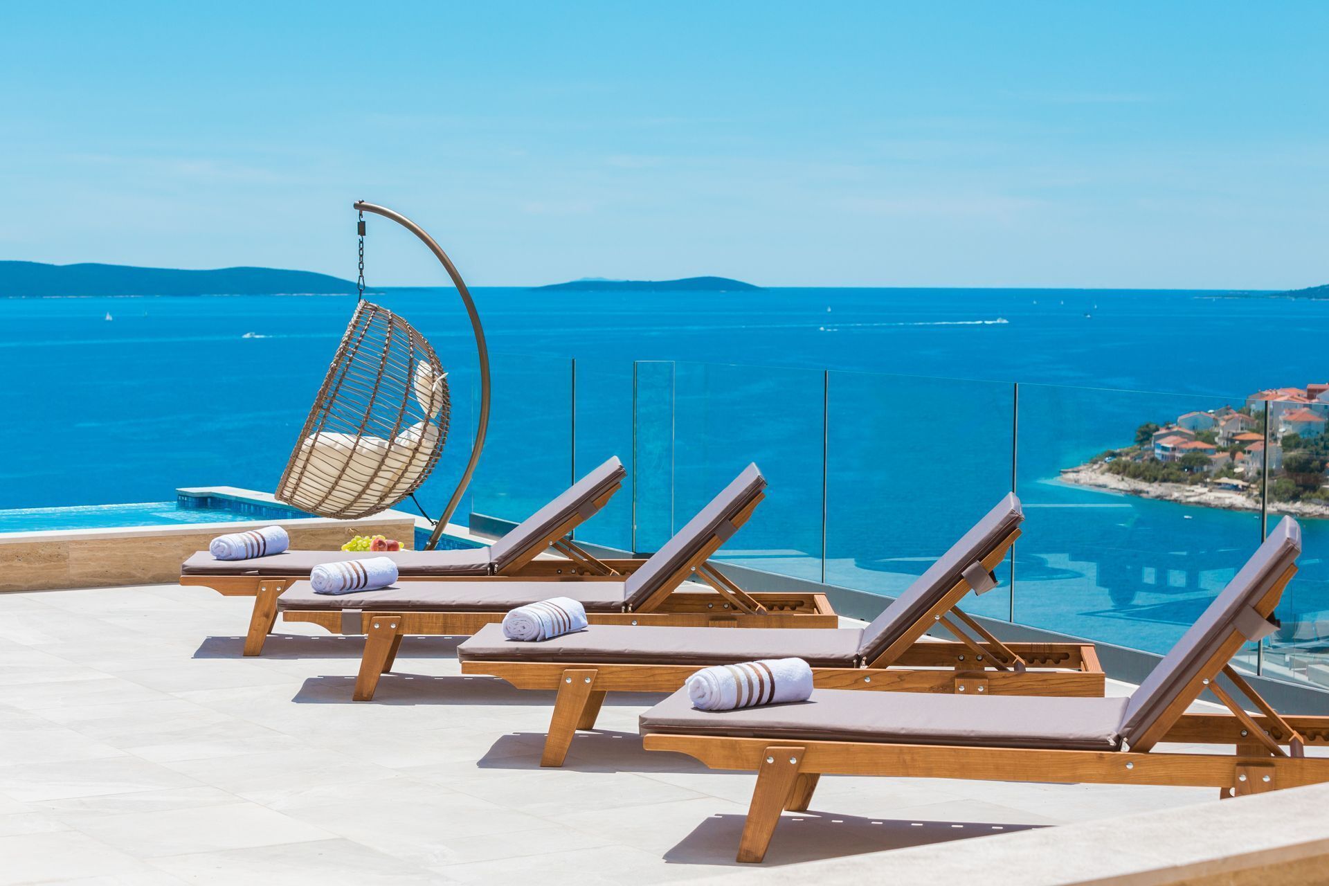 Un espace aménagé au bord de la piscine, avec des chaises longues en bois et un fauteuil suspendu, offrant une vue sur une mer d’un bleu limpide et sur la côte lointaine.