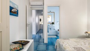 A hotel room with a bed, a bench with cushions, a bathroom with a sink and mirror, and a hallway with a painting. - House with terrace, 80 metres from beach of San Vito lo Capo, Sicily (San Vito Lo Capo)