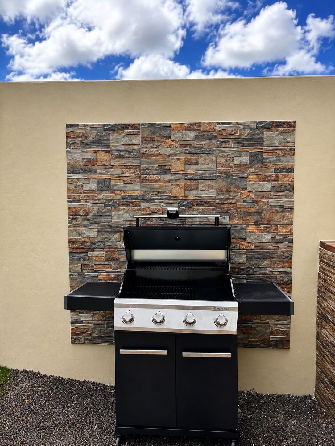 An outdoor grill with a stone wall backdrop, set on a gravel surface.