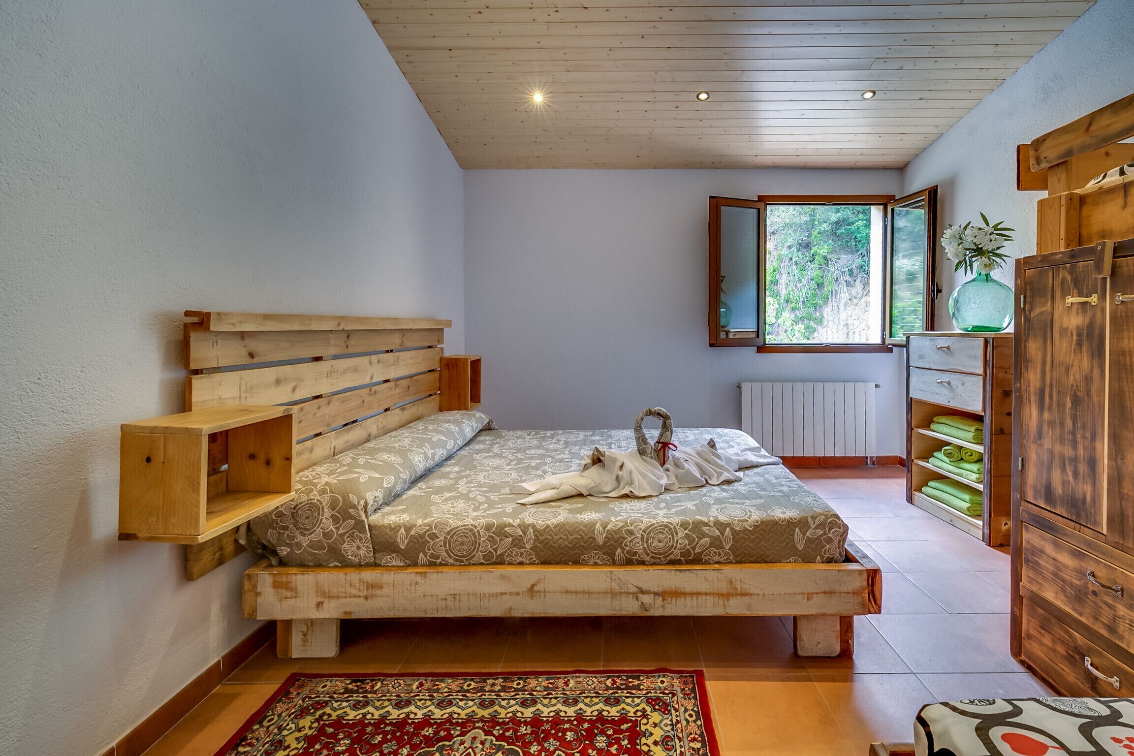 A bedroom with a wooden bed, a large mirror, a window with a view of greenery, and a wooden wardrobe.