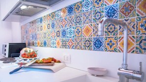 Una cocina con un salpicadero de azulejos coloridos, un microondas, una tabla de cortar con verduras y un fregadero con grifo.