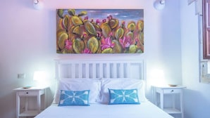 Un dormitorio con una cama blanca, dos almohadas azules con estampado, un cabecero blanco y un cuadro con cactus y flores en la pared.