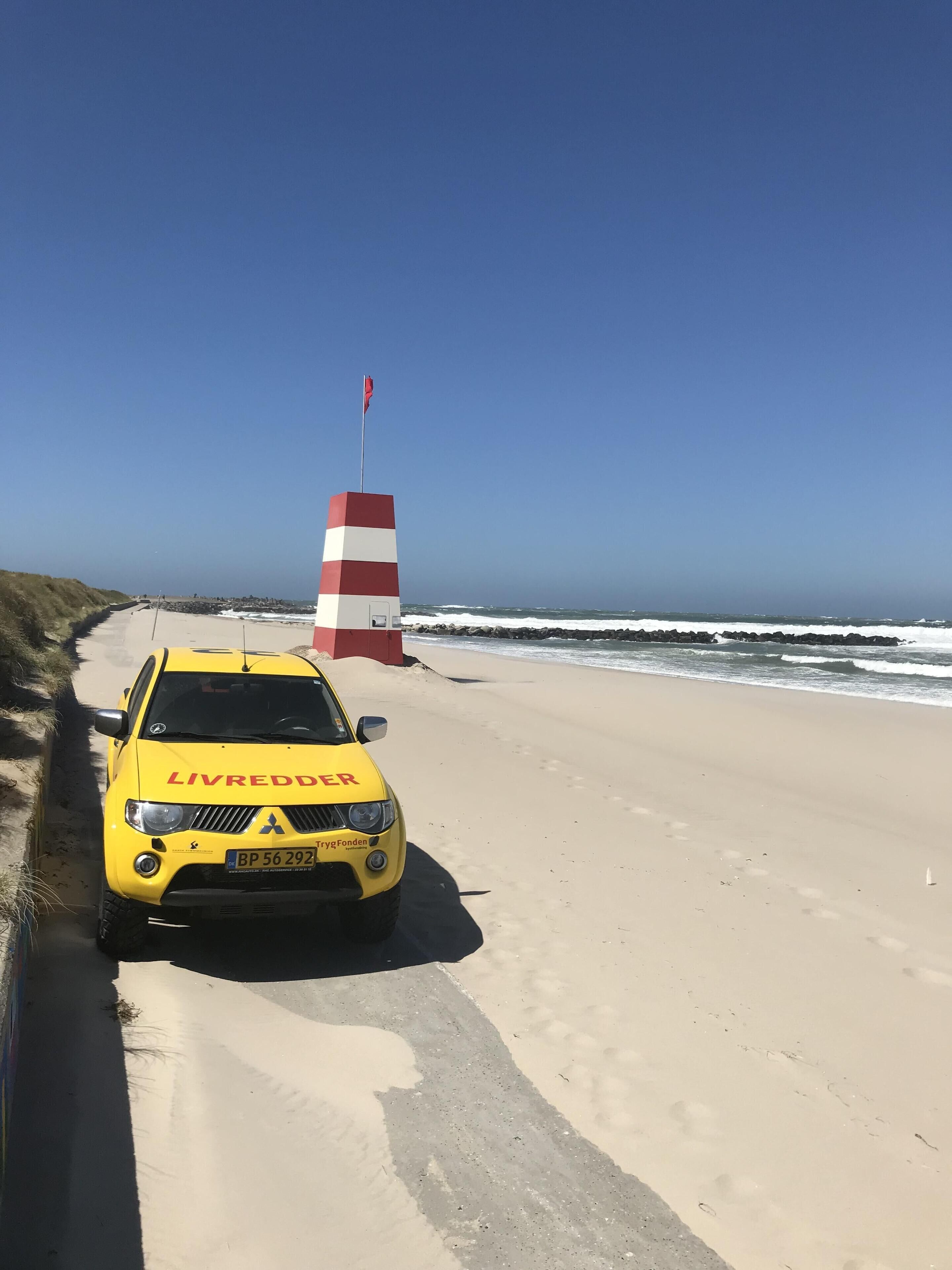 Ein gelbes Fahrzeug mit dem Schriftzug 'LIVREDDER' vorne, geparkt auf einem sandigen Strand mit einem rot-weißen Warnsignal im Hintergrund.