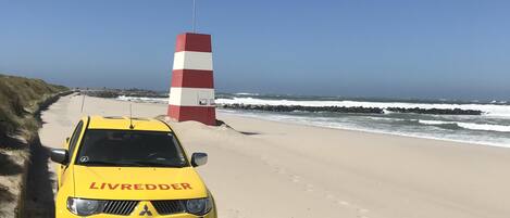 Ein gelbes Fahrzeug mit dem Schriftzug 'LIVREDDER' vorne, geparkt auf einem sandigen Strand mit einem rot-weißen Warnsignal im Hintergrund.