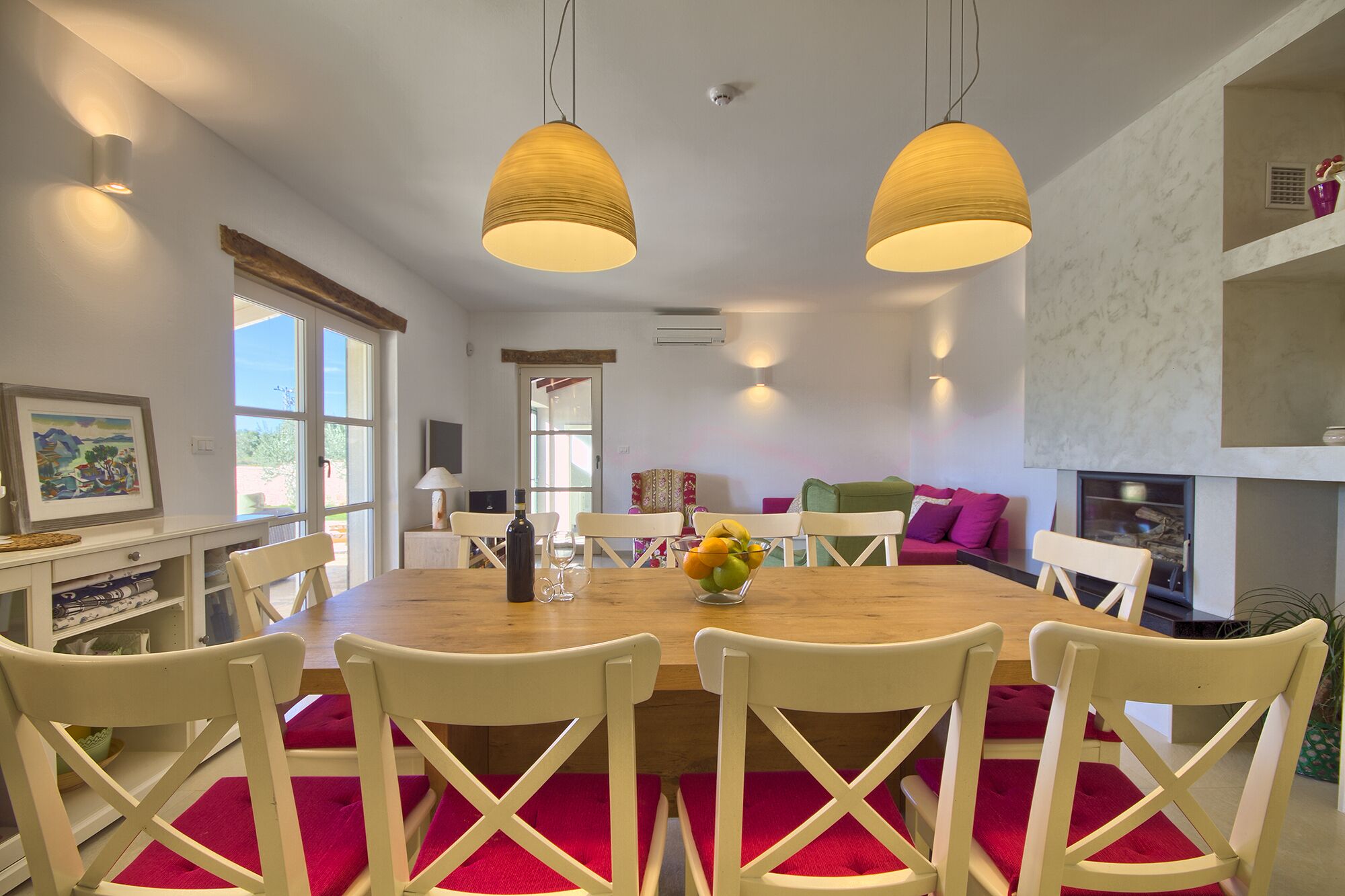 A dining area with a wooden table, white chairs, and a vase of fruit.