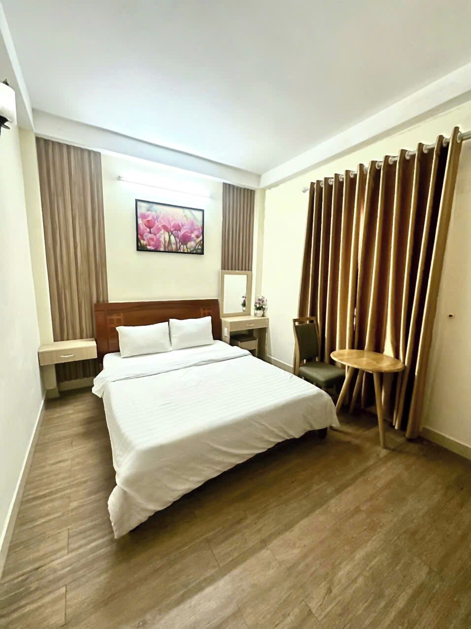 Habitación de hotel con una cama grande, una mesita de noche, una silla, una pequeña mesa, un espejo y cortinas.