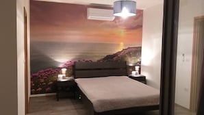 Un dormitorio con cama, lámparas de noche, mesita de noche, un mural en la pared de un atardecer sobre el océano y un equipo de aire acondicionado.