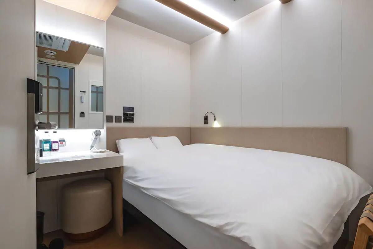 A hotel room with a bed, a bedside table with a lamp, a mirror, and a small trash bin.
