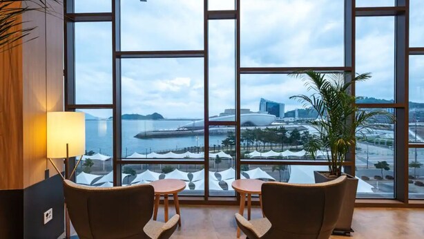 A room with large windows offering a view of a coastal cityscape, including a bridge and buildings. There are two chairs and a small table, with a lamp on the left and a potted plant on the right. - DARAKHYU Yeosu Capsule Hotel by WALKERHILL (Yeosu)