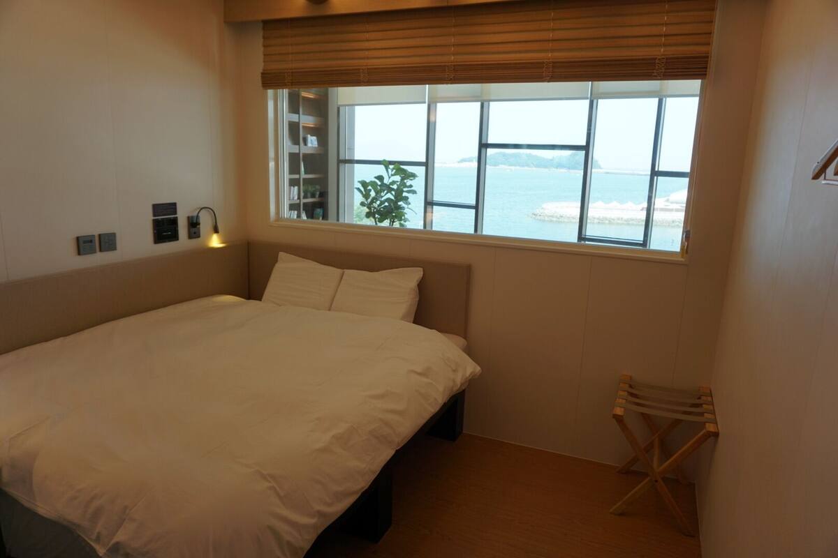 A bedroom with a bed, a window with a view of the sea, a bedside table, and a small plant.