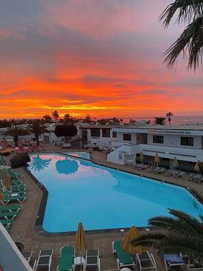 A swimming pool with lounge chairs and umbrellas, surrounded by buildings and palm trees, during sunset. - La Penita - Comfortable apartment with sea view in prime location  (Puerto del Carmen)