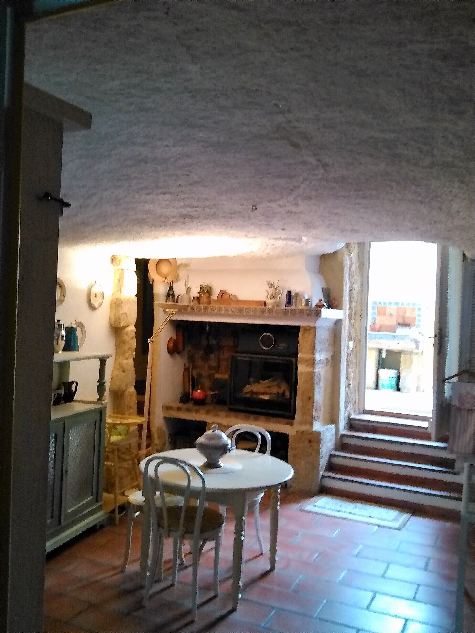 A rustic kitchen with a stone fireplace, a dining table with chairs, and a view of the outside through a doorway.