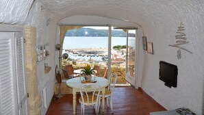A room with a white textured ceiling, a dining area with a table and chairs, a TV mounted on the wall, and a view of a beach and boats through a glass door. - Unusual holiday in a troglodyte house in Saint-Chamas. (Saint-Chamas)