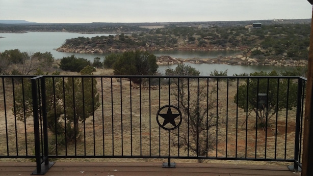 A wooden deck with a metal railing and a star emblem, overlooking a lake and distant buildings. - Beautiful Lakefront Home-Lake Alan Henry (Justiceburg)