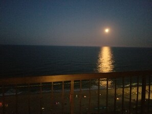 A nighttime view of the ocean with a moon reflection on the water, a wooden railing, and a distant shoreline with lights. - Direct Oceanfront Condo, Amazing Views of Ocean and Bay (Ocean City)