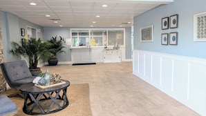 A spacious lobby with a grey armchair, a round table with decorative items, a large potted plant, and a reception desk with a window view. - Direct Oceanfront Condo, Amazing Views of Ocean and Bay (Ocean City)