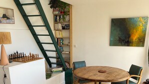 Una sala de estar moderna con piso de madera, una mesa redonda de madera, dos sillas verdes, una estantería, un cuadro y una escalera.
