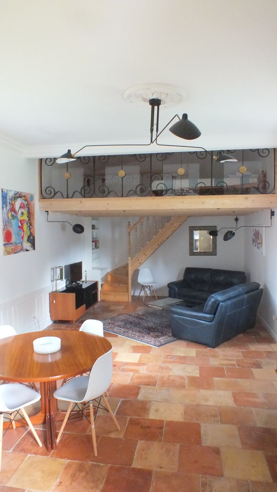 A spacious living area with a wooden staircase, a black leather sofa, a dining table with chairs, and a colorful abstract painting on the wall.