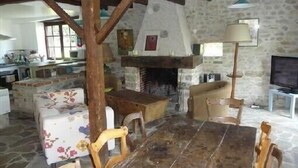 Een landelijke keuken met een stenen muur, houten balken, een open haard, een eettafel met stoelen en een televisie.