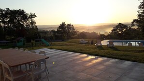 Een terras met een tafel en stoelen, een speeltuin met een glijbaan, en een uitzicht op een meer en bergen tijdens de zonsondergang.