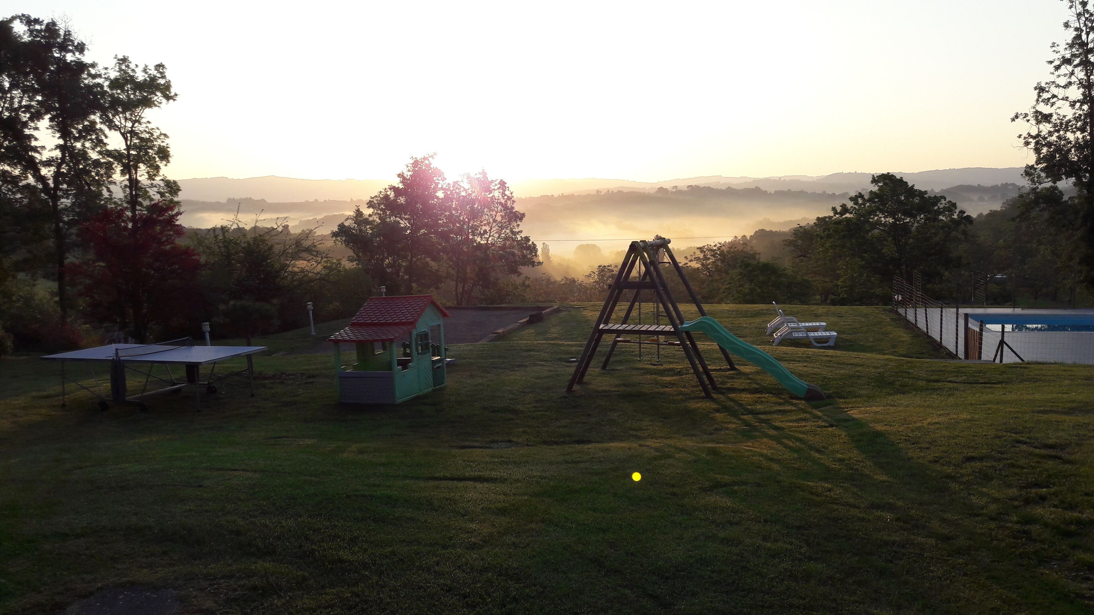 Un parque infantil con columpios y toboganes, rodeado de árboles y vistas a colinas distantes.