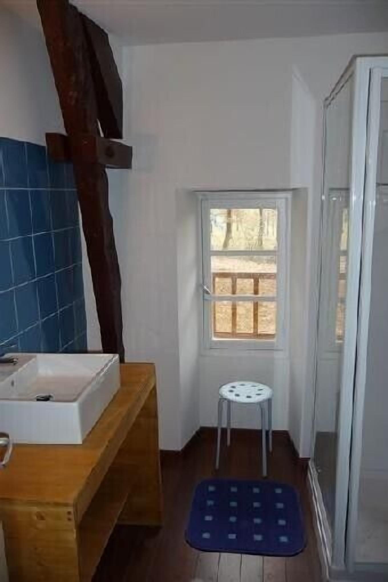 Ein kleines Badezimmer mit einer Holzbank, einem weißen Waschbecken, einem kleinen Hocker und einem Fenster mit Blick ins Freie.