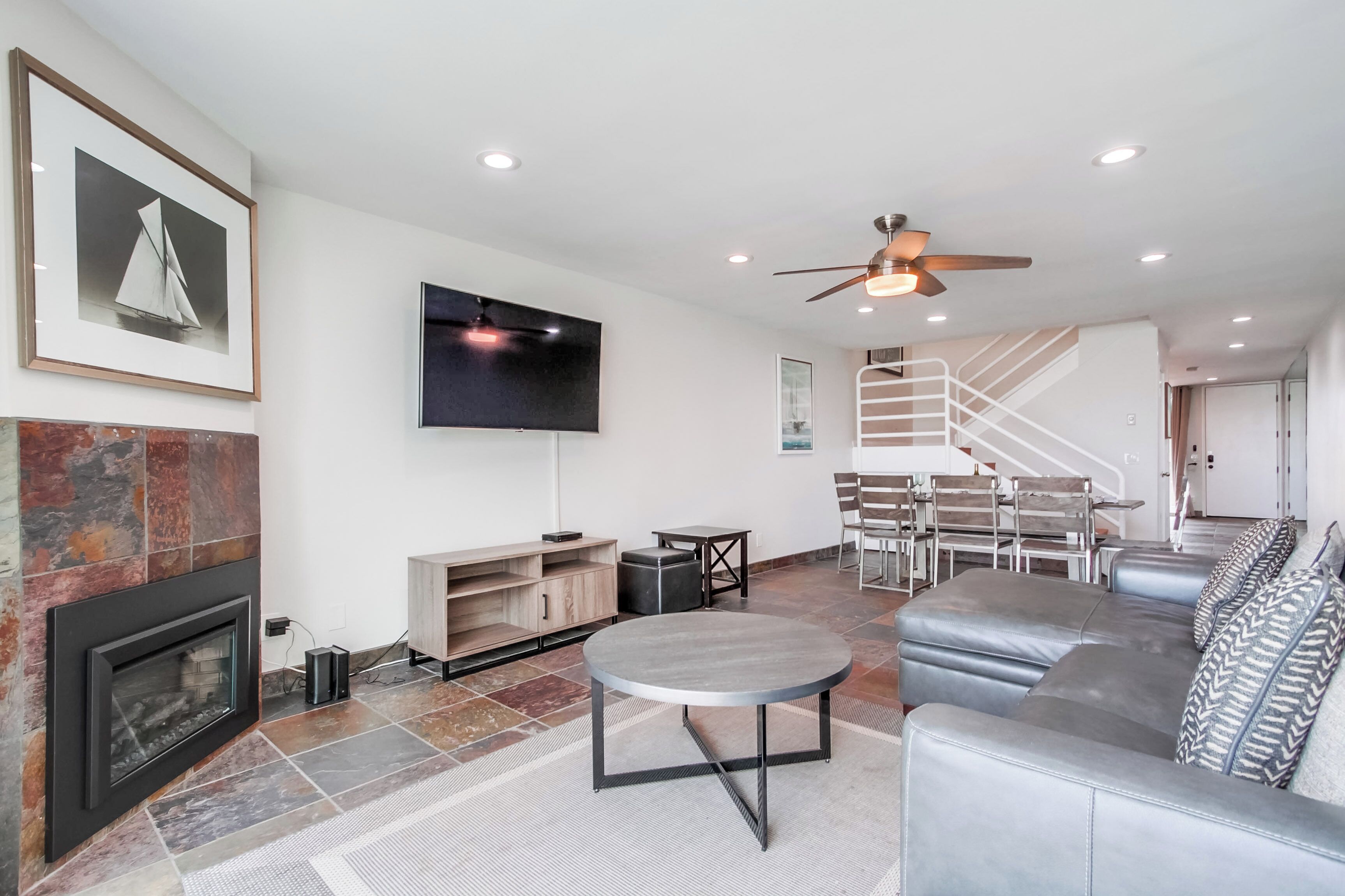A modern living room with a fireplace, a flat-screen TV, a ceiling fan, and a staircase.