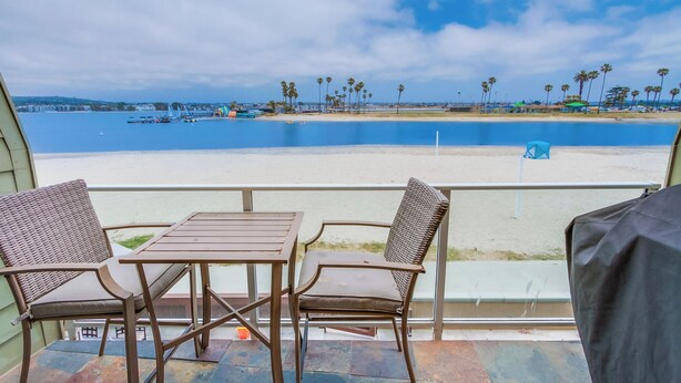 Een balkon met twee rieten stoelen en een tafel, met uitzicht op een strand met palmbomen en een helderblauwe lucht.