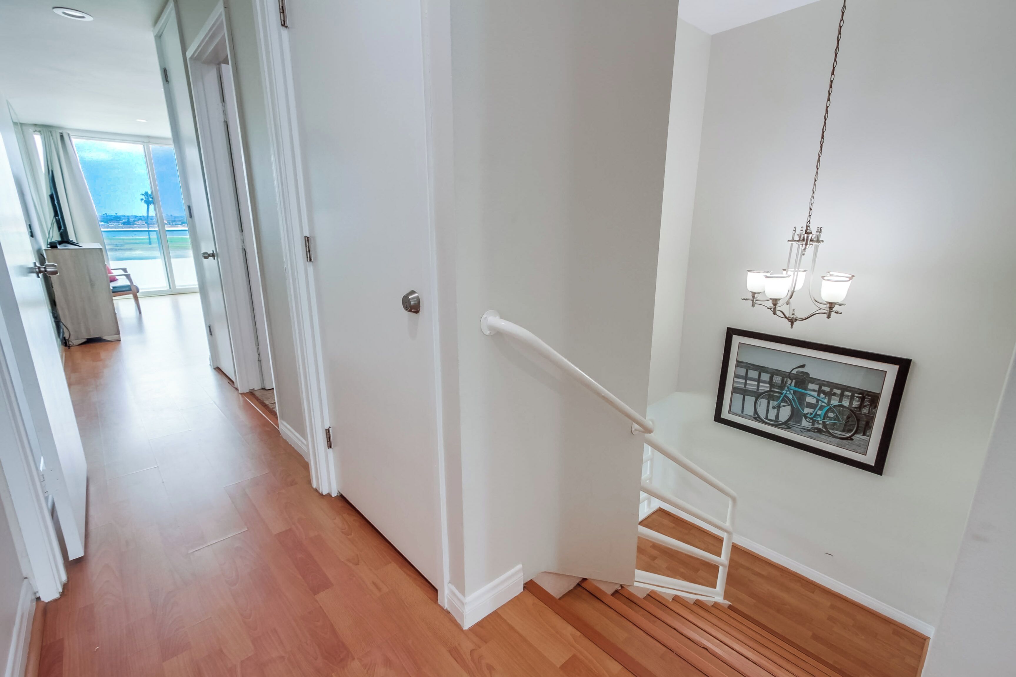 Un pasillo con piso de madera, paredes blancas y una escalera con barandilla blanca. Encima de la escalera, cuelga un cuadro enmarcado.
