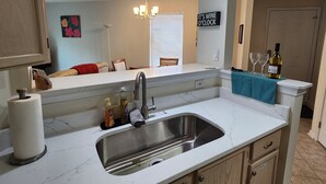 A kitchen with a stainless steel sink, a countertop, and wooden cabinets. There is a sign on the wall that reads "IT'S WINE O'CLOCK".