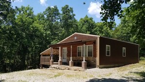 A wooden cabin with a porch, surrounded by trees and a gravel driveway.
