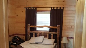 A small, cozy wooden room with a bed, a nightstand, a lamp, and a window with curtains.