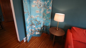 A room with a blue wall, a wooden floor, a red sofa, a round table with a lamp, and a blue folding screen with floral patterns. - West Inn on Weston - Great for Families (Niagara Falls)