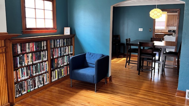 A room with a wooden bookshelf, a blue armchair, a dining table with chairs, and a kitchen area visible through an archway. - West Inn on Weston - Great for Families (Niagara Falls)