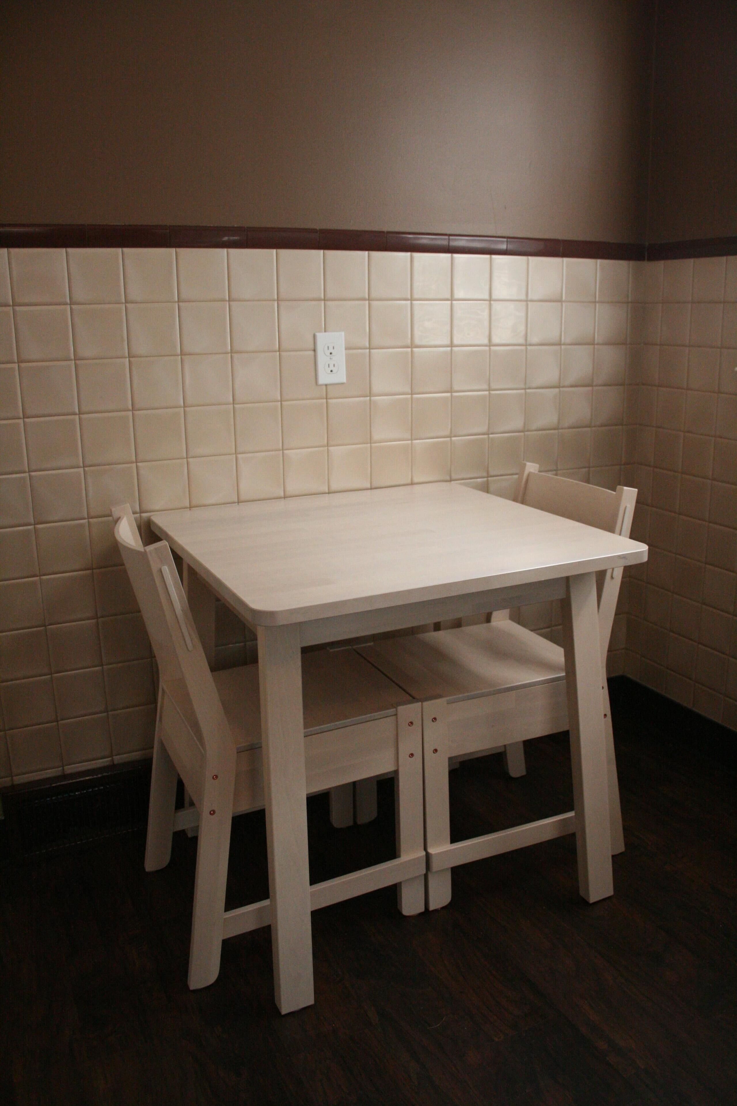 A small, white wooden dining table with four chairs in a corner of a room with tiled walls and a dark wooden floor.
