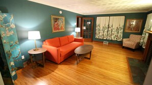 A living room with a wooden floor, a red sofa, a round coffee table, two side tables with lamps, a patterned chair, and a door with curtains. - West Inn on Weston - Great for Families (Niagara Falls)