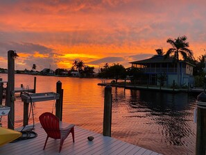 A wooden dock with a red chair and a yellow umbrella, overlooking a calm body of water with a house and palm trees in the background.