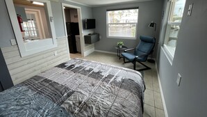 Een kleine kamer met een bed, een stoel, een televisie, een klein tafeltje met een plantje, en een raam met uitzicht op de straat.