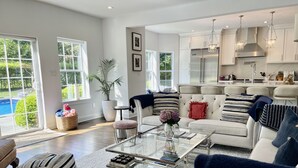 A spacious living room with a large glass coffee table, a white sofa, and a kitchen area with white cabinets and stainless steel appliances.