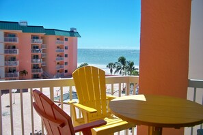 A balcony with yellow chairs and a table, overlooking a beach and a multi-story building with balconies.