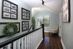A hallway with framed pictures on the walls, a white railing, and a chair in the corner.