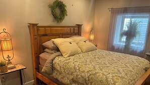 A bedroom with a wooden bed frame, a green patterned bedspread, a wooden nightstand with a lamp, and a window with a view.